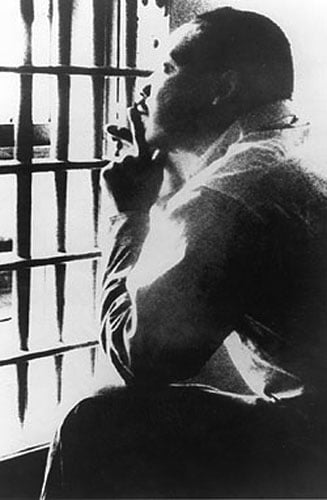 Black and white historical photo: Martin Luther King Jr. looks through the bars of a Burmingham, Alabama, cell in April 1963.