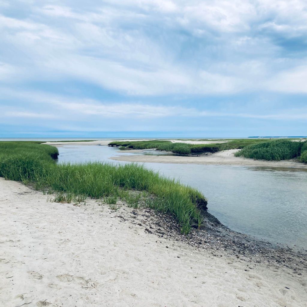 A beautiful beach landscape with white sand, green seagrass, and a blue channel of water under a partly sunny sky.