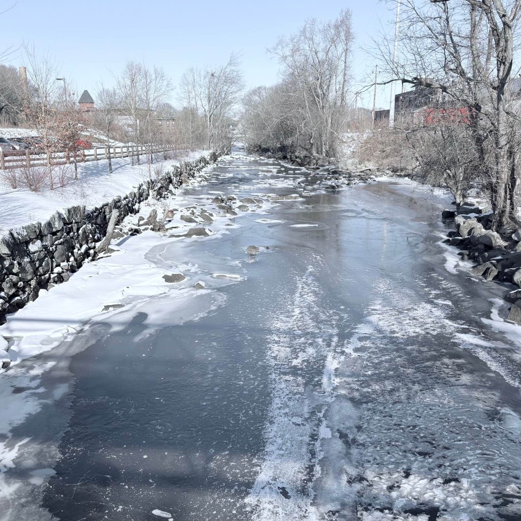 Photograph of a very cold scene: you are standing on a bridge over a small river, which is entirely covered in ice and snow.