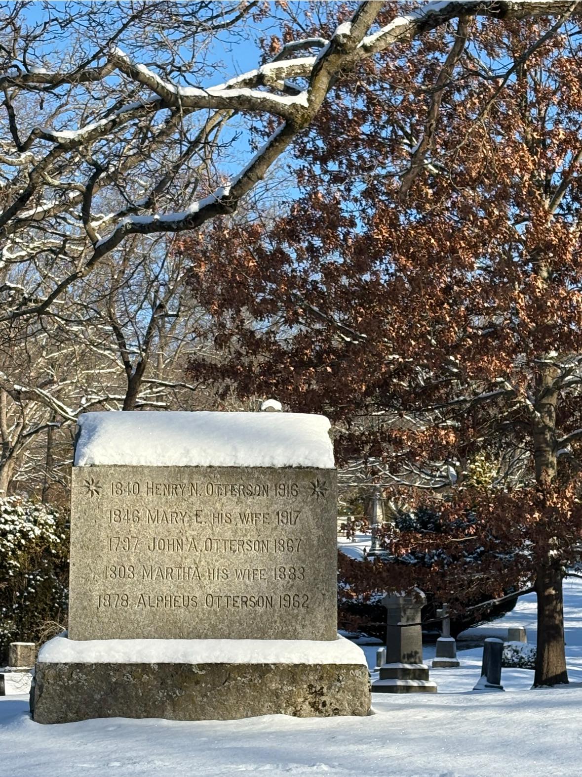 Photograph of a snowy graveyard on a sunny day. We are looking at a granite gravestone with many generation of names engraved on it.