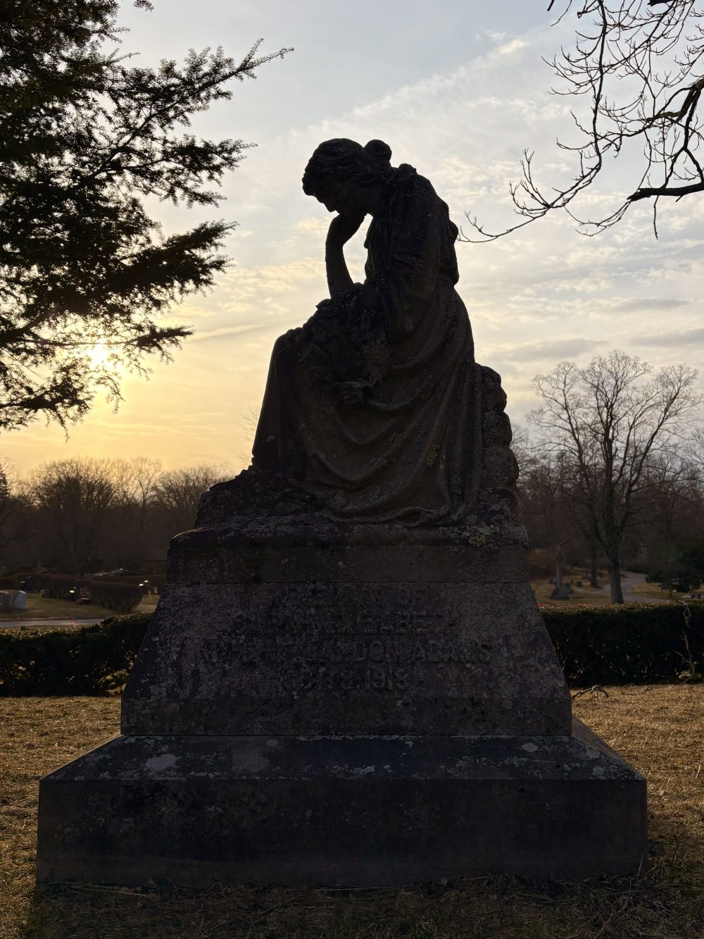 My photo of a memorial in Coffeewalk Cemetery: dawn light is the backdrop for a statue of a seated woman with her chin on her hand, looking down in sadness.