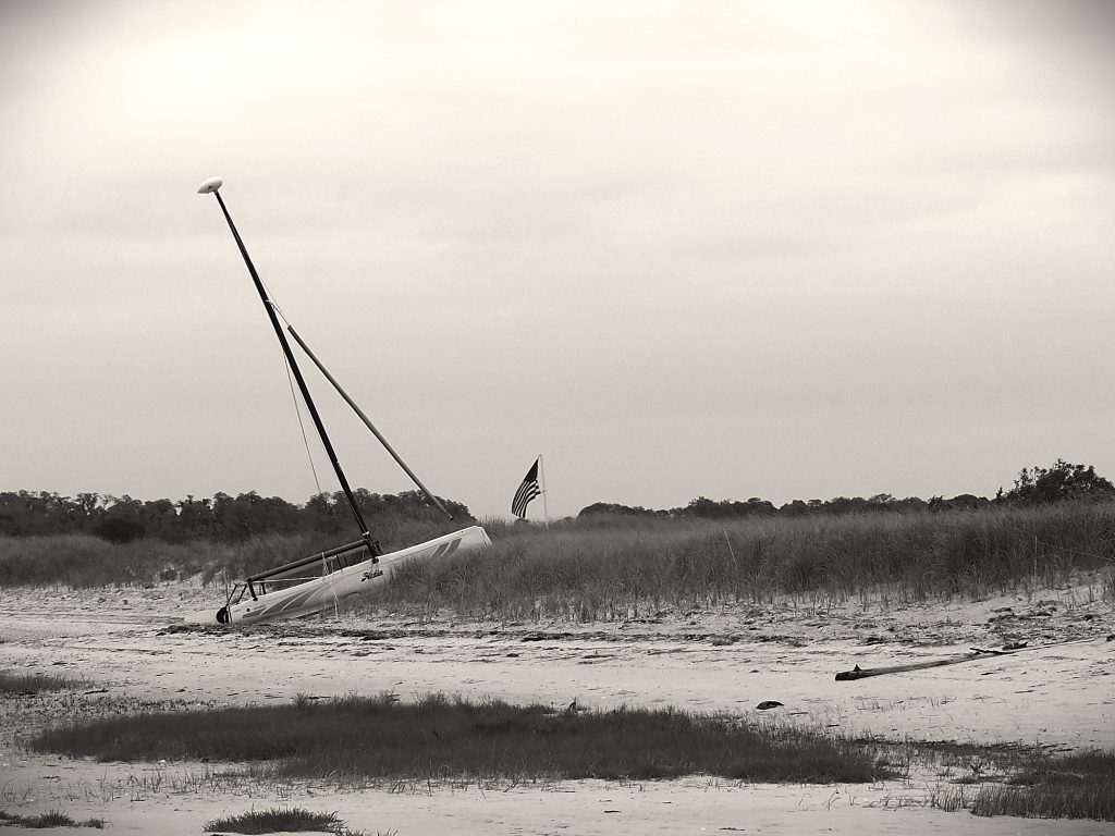 A small sailboat has run aground on a sand dune, pointing towards a drooping American flag. The image is in shadowy black and white.