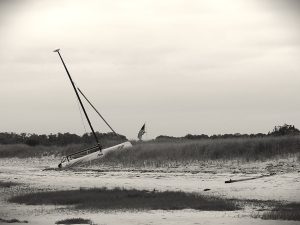 A small sailboat has run aground on a sand dune, pointing towards a drooping American flag. The image is in shadowy black and white.
