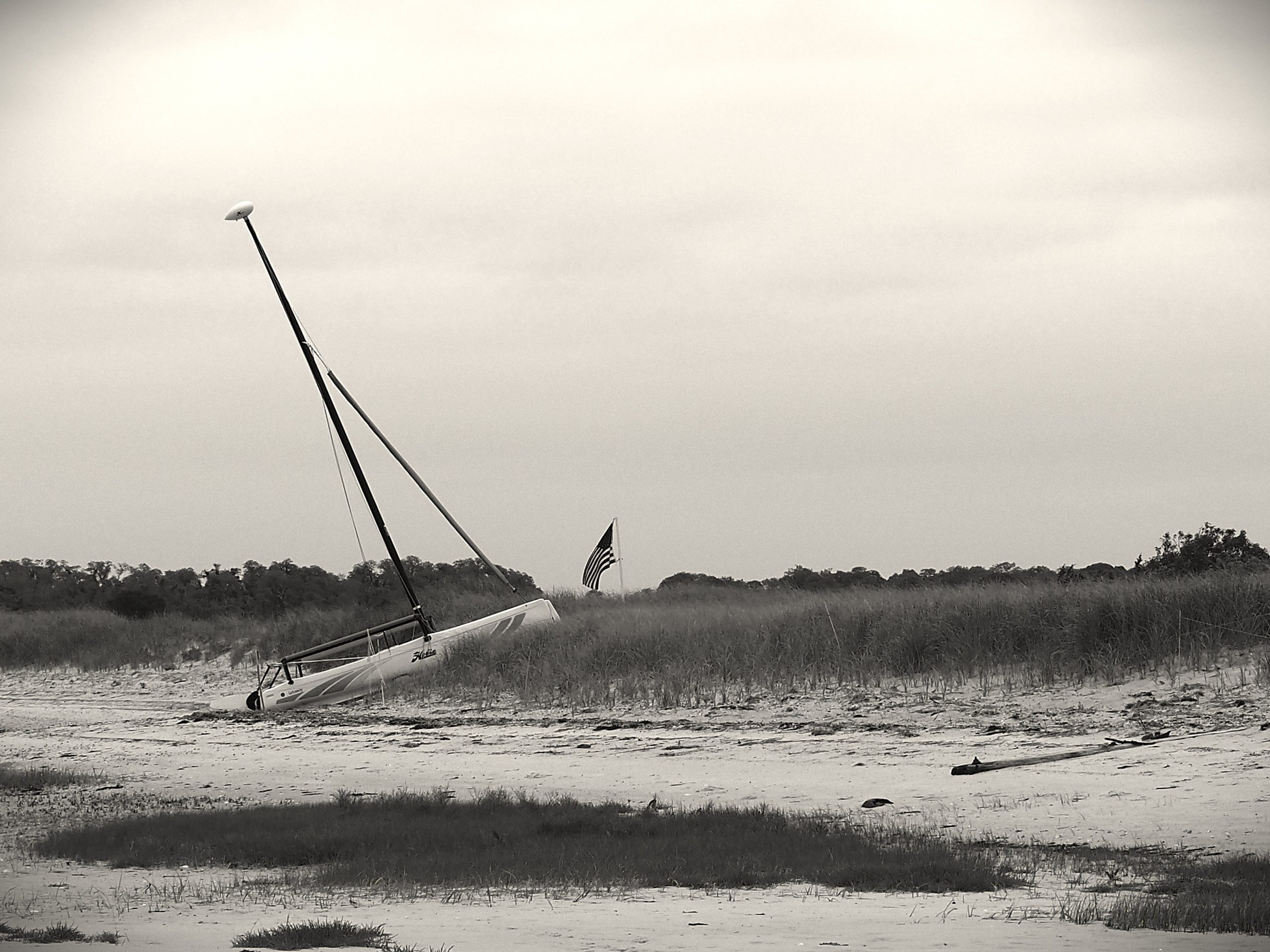 A small sailboat has run aground on a sand dune, pointing towards a drooping American flag. The image is in shadowy black and white.
