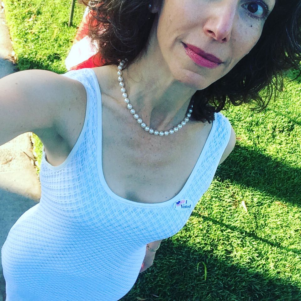 Photo of me leaving my polling place on Nov. 8, 2016. I am wearing a suffragette-white sleeveless dress and I have an "I Voted" sticker over my heart. I'm standing on green grass with an American flag behind me.