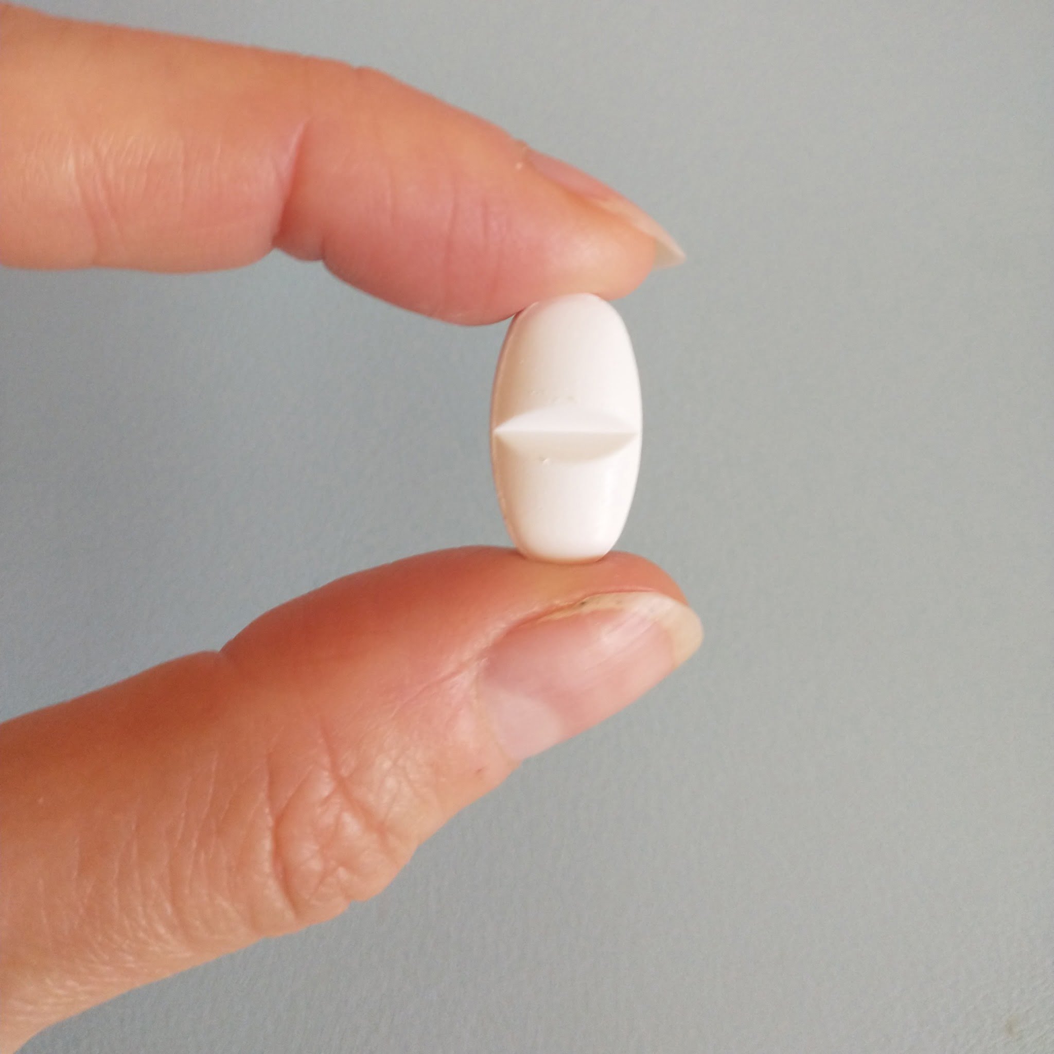 Close-up photo of my index finger and thumb holding a small white pill in front of a light-blue background