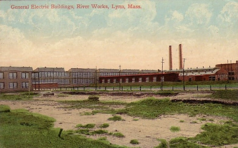 Vintage image of the outside of a factory with a scruffy lawn in foreground, with a caption saying "General Electric Buildings, River Works, Lynn, Mass."