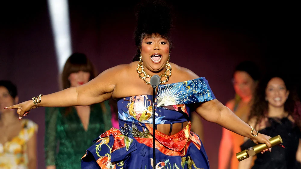 Photo of the performer Lizzo looking beautiful in an evening gown with her arms spread wide as she speaks into a microphone