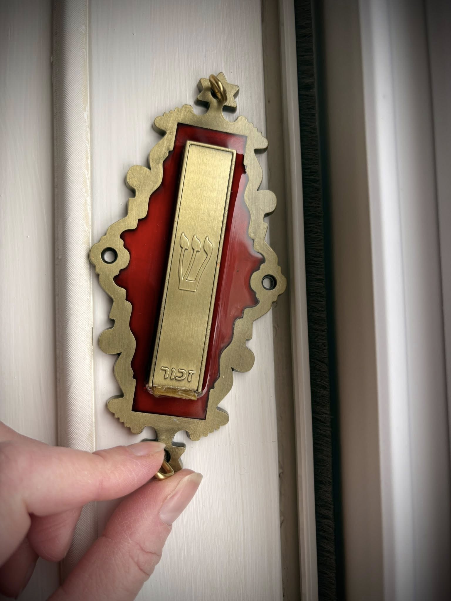 On the white-painted doorway of my home, my fingers are turning the screw to affix a brass-and-red-enamel mezuzah.