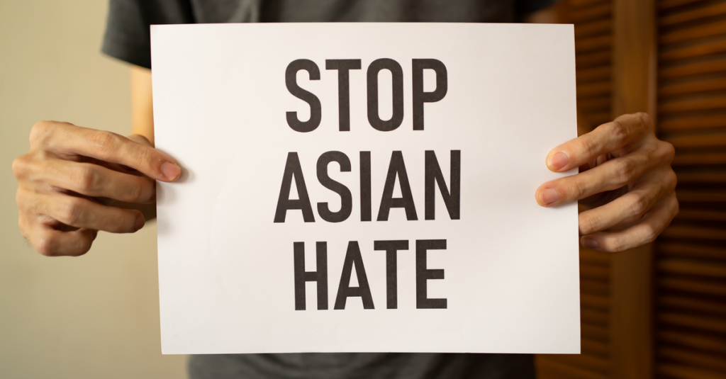 Photo of a man's hands holding a white placard with black all-caps words on it saying "STOP ASIAN HATE"