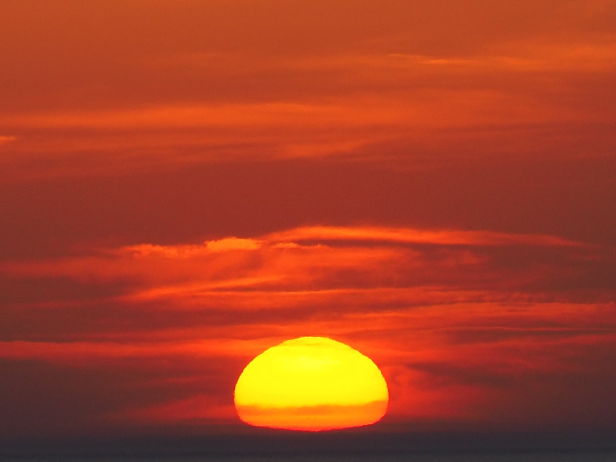 Zoomed-in shot of the setting sun on the horizon, distorted by the atmosphere and surrounded by bright red-orange clouds.