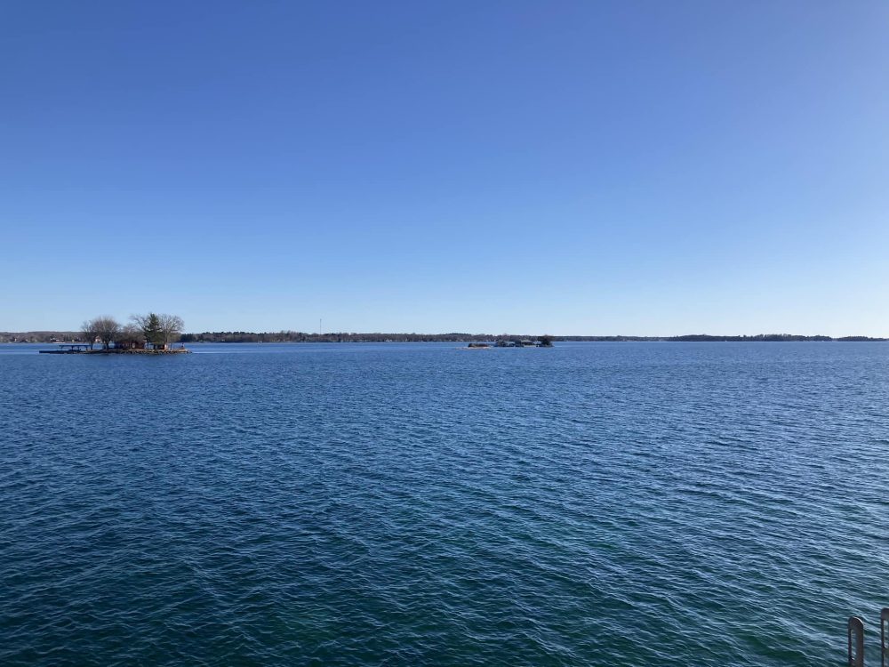 An expanse of deeply blue river under deeply blue sky, interrupted only by a couple of tiny rocky islands poking up between the viewer and a thin line of land that's the faraway opposite riverbank.