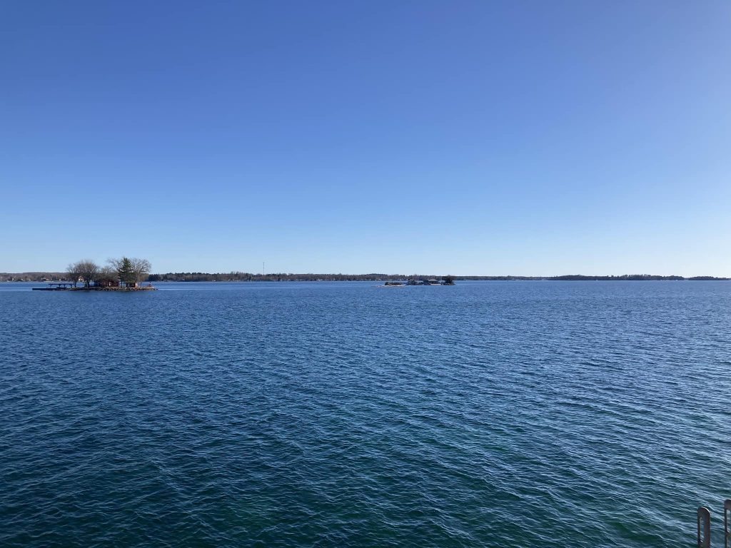 An expanse of deeply blue river under deeply blue sky, interrupted only by a couple of tiny rocky islands poking up between the viewer and a thin line of land that's the faraway opposite riverbank.