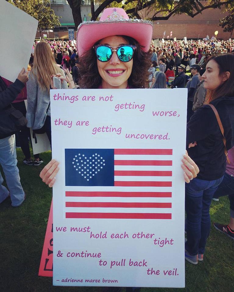 Author holding a protest sign quoting adrienne maree brown's "living through the unveiling"